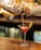 A person is pouring a pink cocktail from a metal shaker into a martini glass with a lime wedge on the rim. The background is softly blurred with warm lighting, suggesting a bar setting. The surface is wooden, and the scene conveys a sense of casual elegance.