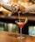 A person is pouring a pink cocktail from a metal shaker into a martini glass with a lime wedge on the rim. The background is softly blurred with warm lighting, suggesting a bar setting. The surface is wooden, and the scene conveys a sense of casual elegance.
