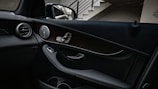 A car interior with focus on the passenger side door panel, featuring a mix of leather and wooden elements with chrome accents. The design includes a speaker, door handle, and control switches. There is a side mirror visible outside, reflecting part of the structure it is parked next to, which includes a staircase.