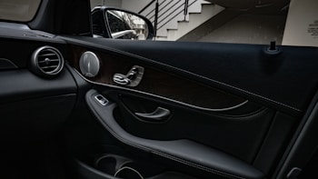 A car interior with focus on the passenger side door panel, featuring a mix of leather and wooden elements with chrome accents. The design includes a speaker, door handle, and control switches. There is a side mirror visible outside, reflecting part of the structure it is parked next to, which includes a staircase.