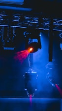 Stage lighting equipment emits vibrant beams of red and blue light, surrounded by a haze that enhances the dramatic effect. The setup includes overhead structures and cables, typical of a professional lighting rig.