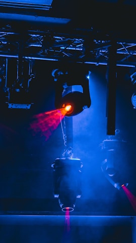 Stage lighting equipment emits vibrant beams of red and blue light, surrounded by a haze that enhances the dramatic effect. The setup includes overhead structures and cables, typical of a professional lighting rig.