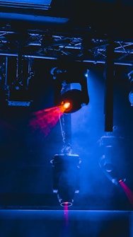 Stage lighting equipment emits vibrant beams of red and blue light, surrounded by a haze that enhances the dramatic effect. The setup includes overhead structures and cables, typical of a professional lighting rig.