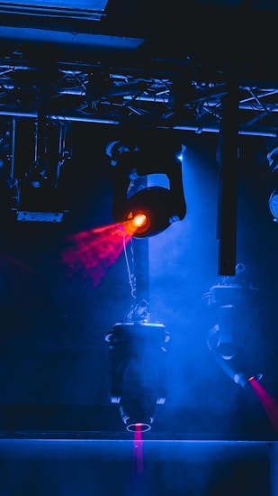 Stage lighting equipment emits vibrant beams of red and blue light, surrounded by a haze that enhances the dramatic effect. The setup includes overhead structures and cables, typical of a professional lighting rig.