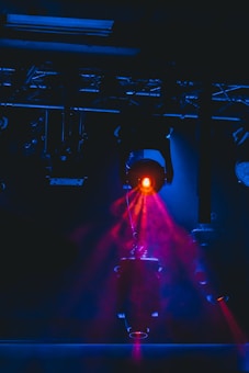 A stage lighting setup featuring a prominent spotlight emitting a bright red beam, creating a vivid contrast against a dark background. The beams of light are interacting with smoke or haze, enhancing the dramatic effect.