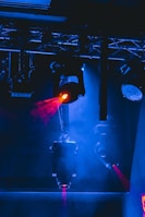 Multiple stage lights suspended from a metal framework illuminate the scene with beams of red light, contrasting against a predominantly blue-lit background. The image creates a dramatic, high-energy ambiance typical of concert or theatrical settings.