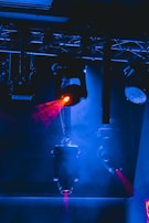 Multiple stage lights suspended from a metal framework illuminate the scene with beams of red light, contrasting against a predominantly blue-lit background. The image creates a dramatic, high-energy ambiance typical of concert or theatrical settings.