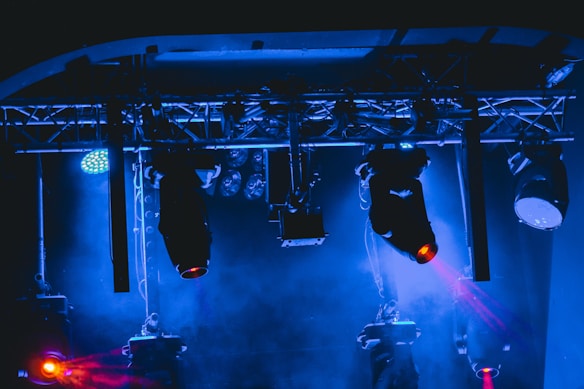 A stage setup featuring various lighting equipment with trusses overhead. The lights emit blue and red colors, creating a dynamic and vibrant ambiance. Smoke or haze adds a dramatic effect to the scene.