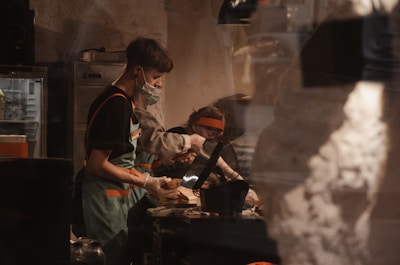 Several people are working in a kitchen or food service environment. One person is wearing a mask and apron, handling food items, while another person is in the background. The atmosphere is dimly lit, with a focus on food preparation.