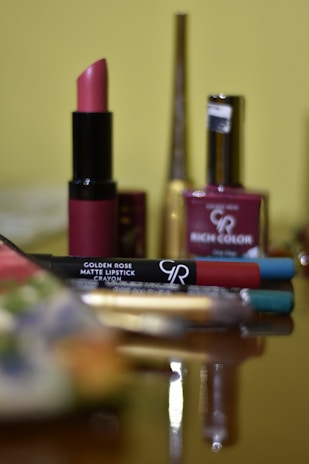 A neatly arranged collection of cosmetics with a prominent rose-colored lipstick in the foreground. Various makeup items, including a nail polish and lip liners, are set against a blurred background.