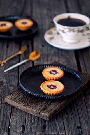 Two small tarts with creamy filling and a berry topping are placed on a dark rustic plate, set on a textured wooden surface. Nearby, a white floral teacup filled with tea sits on a matching saucer. A golden spoon and a silver knife are artfully positioned next to the plate, and another plate with more tarts is visible in the background.