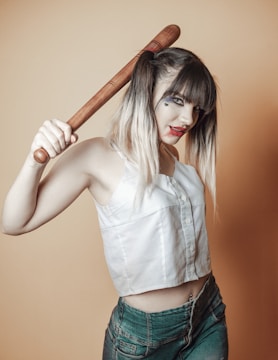 A woman with dark makeup and red lipstick holds a wooden bat over her shoulder. She has blonde and black hair styled in pigtails and is wearing a white sleeveless top and blue jeans. A small heart is drawn near her eye, and she poses with a serious expression against a beige background.