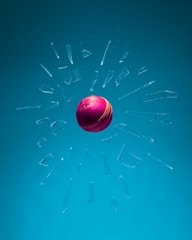A red cricket ball is floating in mid-air surrounded by shattered glass fragments suspended in a blue environment. The ball's stitching and color are prominent, creating a striking contrast against the cool background.