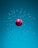 A red cricket ball is floating in mid-air surrounded by shattered glass fragments suspended in a blue environment. The ball's stitching and color are prominent, creating a striking contrast against the cool background.