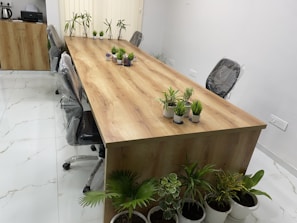 A modern conference room features a long wooden table surrounded by office chairs with protective covering. Small potted plants are arranged across the table, providing a touch of greenery. The floor is tiled in white with subtle marble patterns, and a cabinet with an electric kettle and paper items is seen in the background. Vertical blinds cover the window, and additional potted plants are placed along one side.