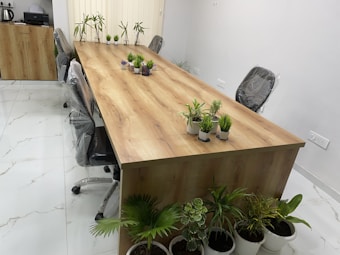 A modern conference room features a long wooden table surrounded by office chairs with protective covering. Small potted plants are arranged across the table, providing a touch of greenery. The floor is tiled in white with subtle marble patterns, and a cabinet with an electric kettle and paper items is seen in the background. Vertical blinds cover the window, and additional potted plants are placed along one side.