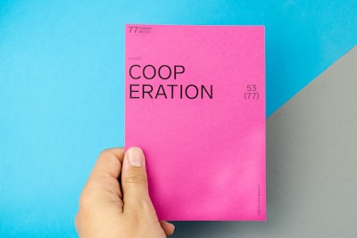 A hand holding a bright pink card with the word 'COOPERATION' printed in bold black letters. The card includes additional text, such as '(social)' and numbers '53 (77)'. The background features a light blue and gray diagonal pattern.