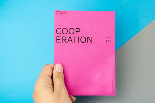 A hand holding a bright pink card with the word 'COOPERATION' printed in bold black letters. The card includes additional text, such as '(social)' and numbers '53 (77)'. The background features a light blue and gray diagonal pattern.