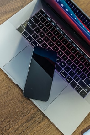 A sleek smartphone is placed on the touchpad of a laptop with a colorful backlit keyboard. The laptop rests on a wooden surface, and the screen displays a gradient of colors.