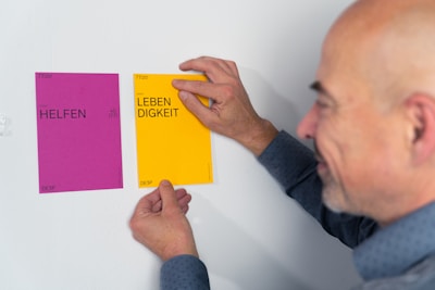 A person is hanging two colorful cards on a white wall. The left card is purple and has the word 'HELFEN' written on it, while the right card is yellow with the word 'LEBENDIGKEIT'. The individual appears to be arranging the cards carefully.