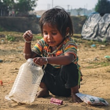 A young child with dark hair and wearing a striped shirt sits on sandy ground. The child is playing with a large, empty plastic jar, appearing curious and engaged. In the background, there is a blurred view of some greenery and a tarp-covered structure, suggesting an outdoor setting.