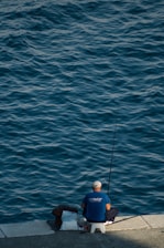 A person sits on a stool by the water's edge, holding a fishing rod. The calm water reflects shades of blue and gives a tranquil atmosphere. The individual wears a blue shirt and a white cap, surrounded by a few bags placed on the ground.