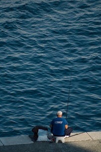 A person sits on a stool by the water's edge, holding a fishing rod. The calm water reflects shades of blue and gives a tranquil atmosphere. The individual wears a blue shirt and a white cap, surrounded by a few bags placed on the ground.