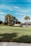 A serene golf course green surrounded by palm trees and a few other trees, with a clubhouse or residential building in the background. The sky is clear with a few clouds, and the overall scene appears calm and peaceful.