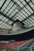 A large, modern stadium with a vast, intricate glass roof structure. Rows of red and green seats are arranged in tiers, bordered by concrete and metal railings. A large cylindrical structure is located in the background, adding to the architectural complexity.