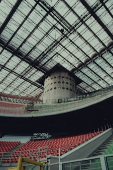 A large, modern stadium with a vast, intricate glass roof structure. Rows of red and green seats are arranged in tiers, bordered by concrete and metal railings. A large cylindrical structure is located in the background, adding to the architectural complexity.