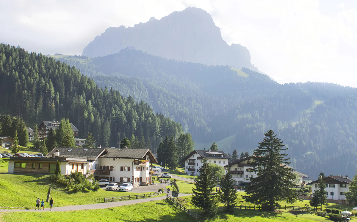 Hotels and chalets on a meadow at Dantercepies above Selva, with pine forest and a hazy rock massif behind