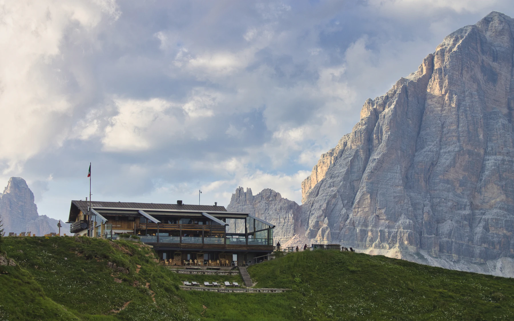Photograph of A timber mountain refuge stands on green grass beneath an immense Dolomites limestone wall.