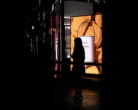 A silhouette of a person is seen in profile against a brightly lit advertisement featuring a Chanel No. 5 product. The background includes various reflections and colorful lights, creating a contrast between the dark and illuminated areas.