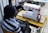 A person with braided hair and wearing a striped shirt is sitting at a wooden table operating a white and pink sewing machine. There is a spool of white thread and a black remote control nearby. Another person is in the background using a different machine.