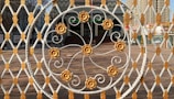 An intricately designed metal gate with a circular pattern featuring floral motifs in gold and white is in the foreground. Behind the gate, a building with a grand staircase and an entrance can be seen. The architecture includes decorative elements and multiple stories with windows, set against an urban backdrop with tall buildings.