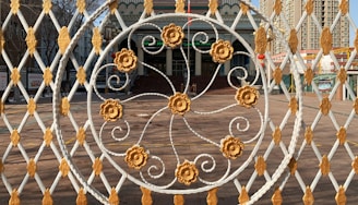 An intricately designed metal gate with a circular pattern featuring floral motifs in gold and white is in the foreground. Behind the gate, a building with a grand staircase and an entrance can be seen. The architecture includes decorative elements and multiple stories with windows, set against an urban backdrop with tall buildings.