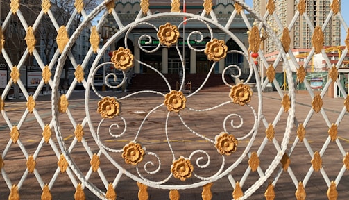 An intricately designed metal gate with a circular pattern featuring floral motifs in gold and white is in the foreground. Behind the gate, a building with a grand staircase and an entrance can be seen. The architecture includes decorative elements and multiple stories with windows, set against an urban backdrop with tall buildings.