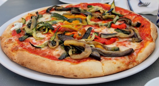 A freshly baked pizza topped with colorful roasted vegetables including red bell peppers, zucchini, and eggplant. The pizza rests on a white plate placed on a table, and the crust appears crispy and golden brown.