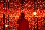 A person is standing in front of a backdrop filled with numerous glowing red lights. The lights create a mesmerizing and immersive environment. The person is holding a camera, capturing the scene or possibly taking a selfie.