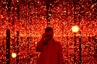 A person is standing in front of a backdrop filled with numerous glowing red lights. The lights create a mesmerizing and immersive environment. The person is holding a camera, capturing the scene or possibly taking a selfie.