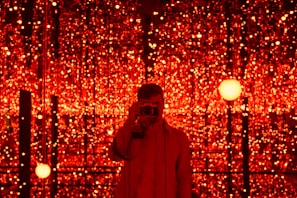A person is standing in front of a backdrop filled with numerous glowing red lights. The lights create a mesmerizing and immersive environment. The person is holding a camera, capturing the scene or possibly taking a selfie.