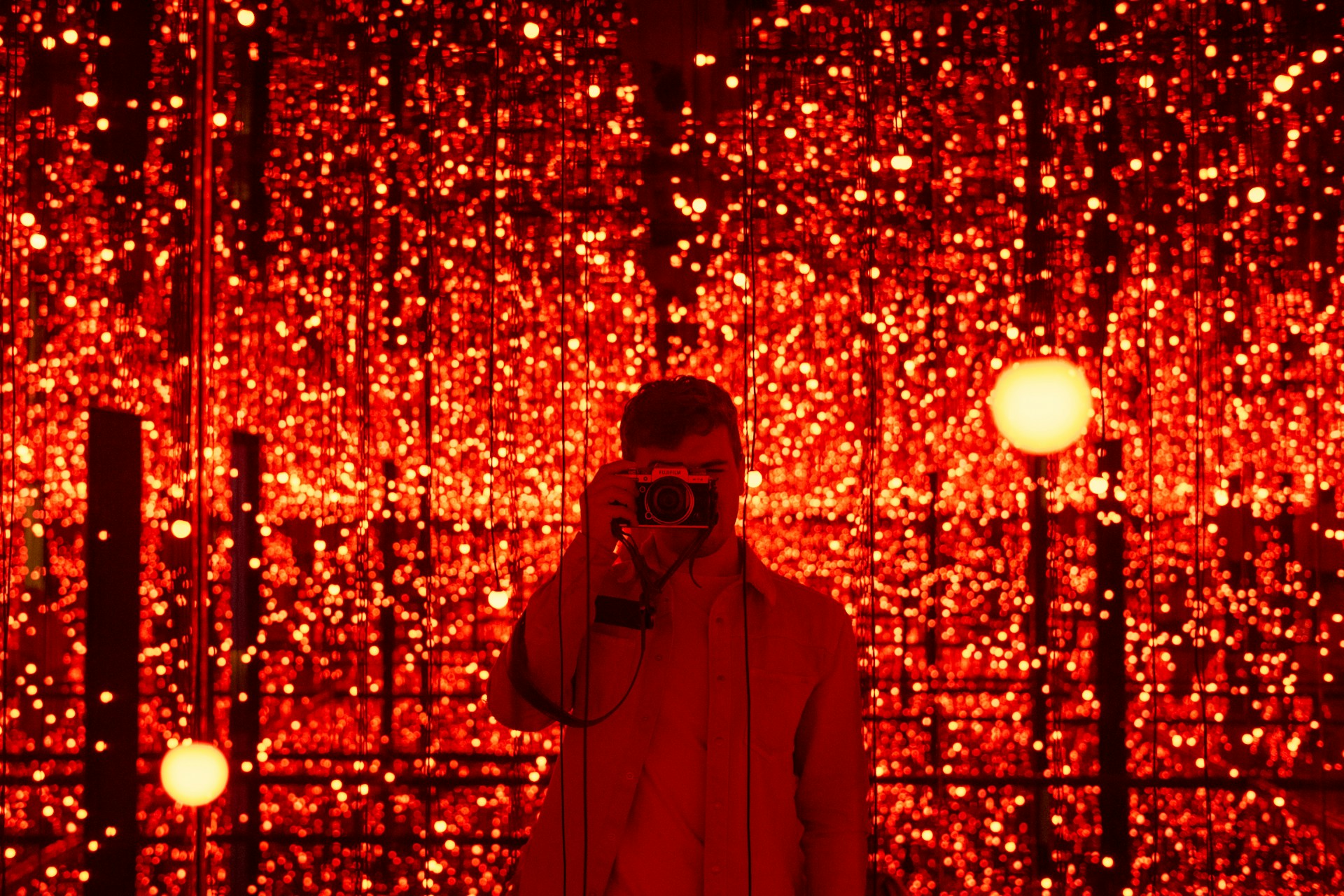 A person is standing in front of a backdrop filled with numerous glowing red lights. The lights create a mesmerizing and immersive environment. The person is holding a camera, capturing the scene or possibly taking a selfie.