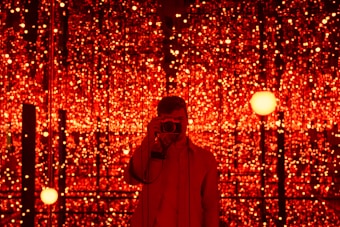 A person is standing in front of a backdrop filled with numerous glowing red lights. The lights create a mesmerizing and immersive environment. The person is holding a camera, capturing the scene or possibly taking a selfie.