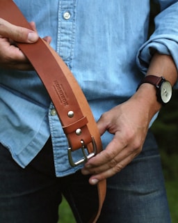 A person holding a brown leather belt with a metal buckle, wearing a light blue denim shirt and dark jeans. On their left wrist, there's a watch with a black strap. The background is blurred, and the image is focused on the belt and the person's hands.