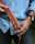 A person holding a brown leather belt with a metal buckle, wearing a light blue denim shirt and dark jeans. On their left wrist, there's a watch with a black strap. The background is blurred, and the image is focused on the belt and the person's hands.