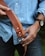 A person holding a brown leather belt with a metal buckle, wearing a light blue denim shirt and dark jeans. On their left wrist, there's a watch with a black strap. The background is blurred, and the image is focused on the belt and the person's hands.