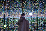 A person stands in front of a backdrop filled with an array of colorful, twinkling lights, capturing a photograph. The background creates an immersive, almost infinite illusion with its reflections.