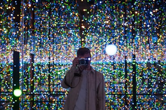 A person stands in front of a backdrop filled with an array of colorful, twinkling lights, capturing a photograph. The background creates an immersive, almost infinite illusion with its reflections.