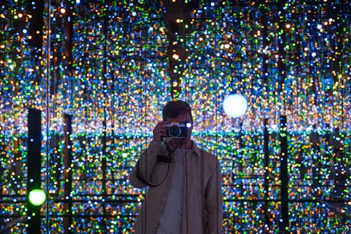 A person stands in front of a backdrop filled with an array of colorful, twinkling lights, capturing a photograph. The background creates an immersive, almost infinite illusion with its reflections.