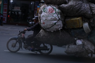 A motorcycle carries a large, overloaded pile of burlap sacks and various items tightly bound together with cords. The rider, wearing a helmet, navigates through an urban street. The scene is slightly dim, indicating possible dusk or evening lighting.